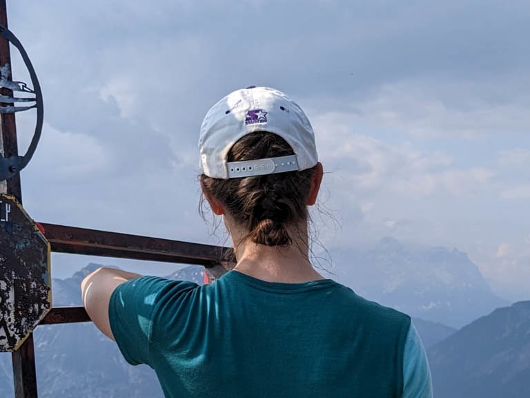 Am metallenen Gipfelkreuz zeigt Bergwanderführerin Charlotte Malycha mit ausgestrecktem Arm in die alpine Bergkette.
