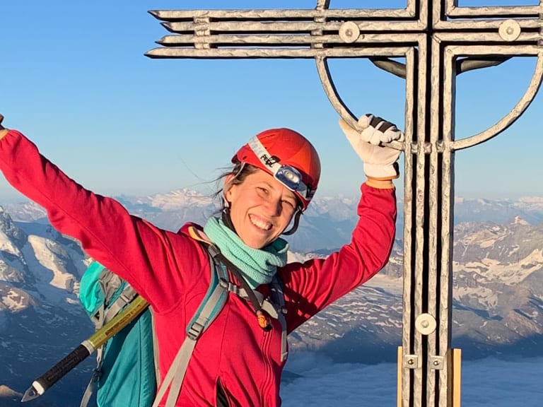 Bergwanderführerin Charlotte Malycha streckt beide Arme jubelnd am hölzernen Gipfelkreuz hoch, trägt rote Jacke und Eispickel.