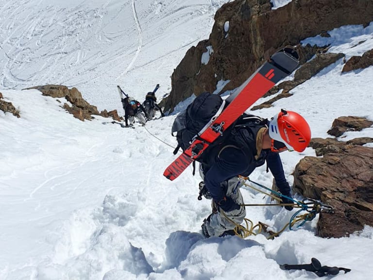 Bergführer Tim Lenz steigt mit Eisgeräten durch steilen, verschneiten Felshang, Skier am Rucksack befestigt.
