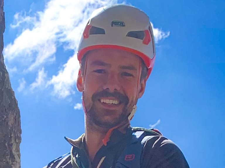 Lächelndes Portrait von Bergführer Tim Lenz in blauem Hemd mit weißem Kletterhelm vor Felswand.