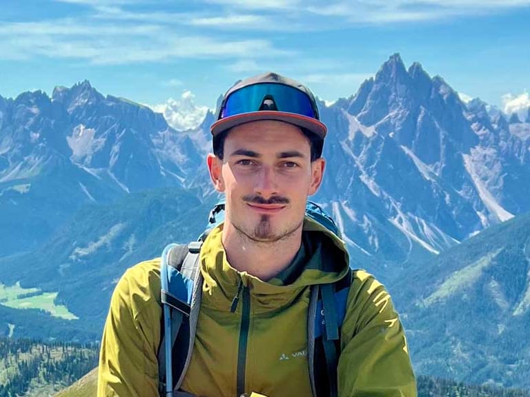 Bergwanderführer Tobias Storz sitzt mit gelbem Cap und Softshelljacke auf steinigem Berghang vor Berglandschaft.
