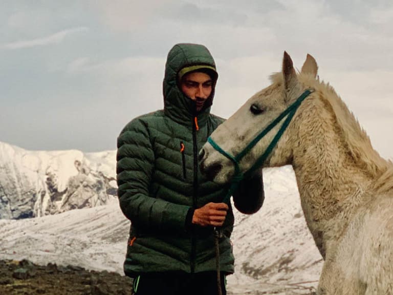 In grüner Winterjacke steht Bergwanderführer Tobias Storz neben hellem Pferd in verschneiter Hochgebirgslandschaft.