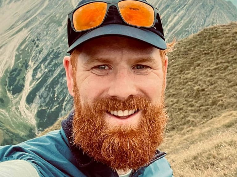 Selfie von Bergwanderführer Dennis Enders mit orangener Gletscherbrille vor alpiner Bergkulisse mit Gipfelkreuz.