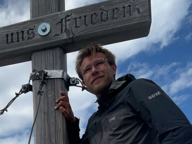 Bergwanderführer Johannes Geipel steht am Holzkreuz eines Berggipfels und berührt den Kreuzpfosten mit der rechten Hand.
