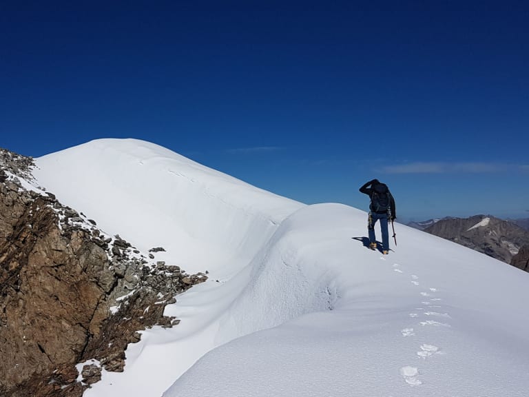 Bergwanderführer Johannes Geipel steht mit Steigeisen auf verschneitem Grat zwischen steiler Felswand und weißer Firnfläche bei klarem Himmel.