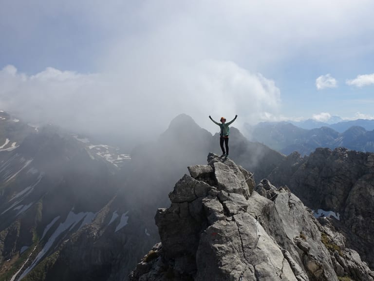 Mit erhobenen Armen jubelt Bergwanderführerin Marie Nimsgern auf einem hellen Kalksteingipfel vor weitem Alpenpanorama.