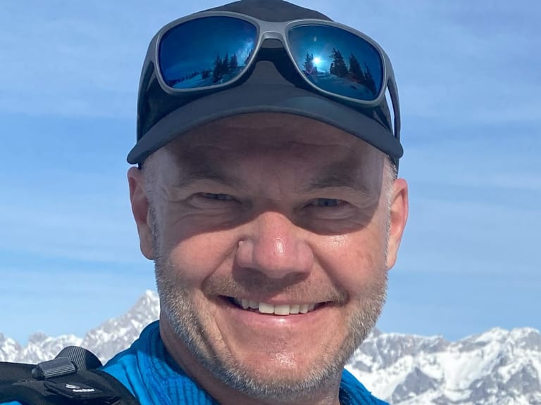 Portrait von Bergwanderführer Gerald Römpp mit dunkler Mütze und Sonnenbrille vor verschneiter Bergkulisse.