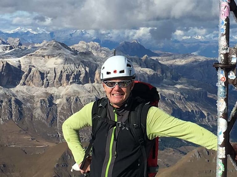 Bergwanderführer Gerald Römpp mit weißem Helm und Klettergurt steht am metallenen Gipfelkreuz vor weitem Bergpanorama.