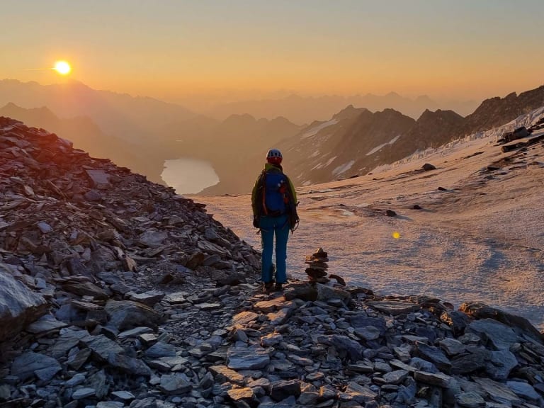 Bergwanderführerin Sophie Sasse steht in blauer Jacke auf steinigem Bergplateau und blickt bei Sonnenaufgang über mehrere Bergketten.
