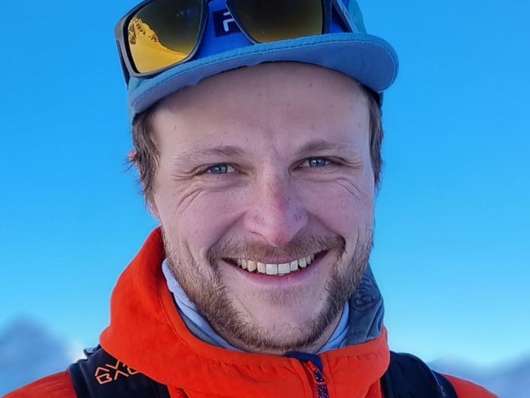 Bergführer Tobias Kostenbader lächelt vor verschneiter Bergkulisse, trägt orange-schwarze Winterjacke und blaue Mütze.