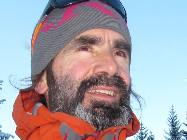 Portrait von Bergwanderführer Karsten Clauß mit grauer Strickmütze und orange-grauer Winterjacke vor verschneiten Nadelbäumen.