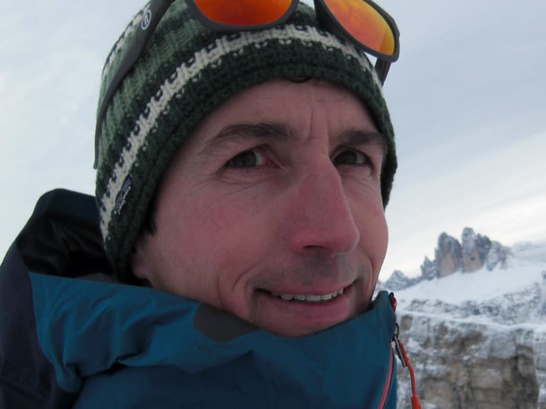 Portrait der Bergführerin Matthias Larcher mit grüner Wollmütze und Bergsteigerbrille vor schneebedeckten Gipfeln im Winter.