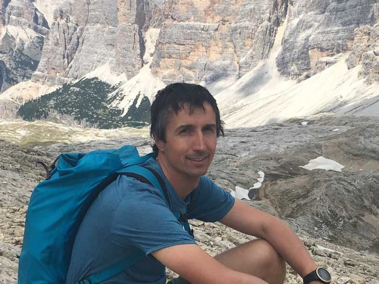 In blauem T-Shirt und grünen Shorts sitzt Bergführerin Matthias Larcher entspannt auf Felsvorsprung vor alpiner Karstlandschaft.