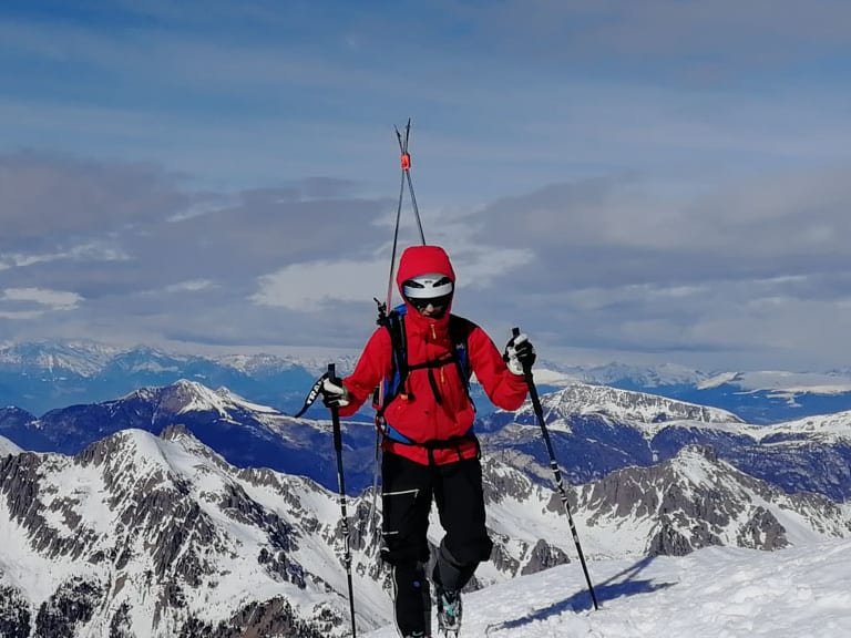 Auf verschneitem Gipfel steht Bergführer Cesare Lotti mit Tourenskiern vor Alpenpanorama.