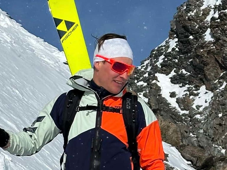 Bergführer Robert Kadner steht mit Skistöcken und gelbem Ski am steilen Schneehang vor massiver Felswand bei Sonnenschein.