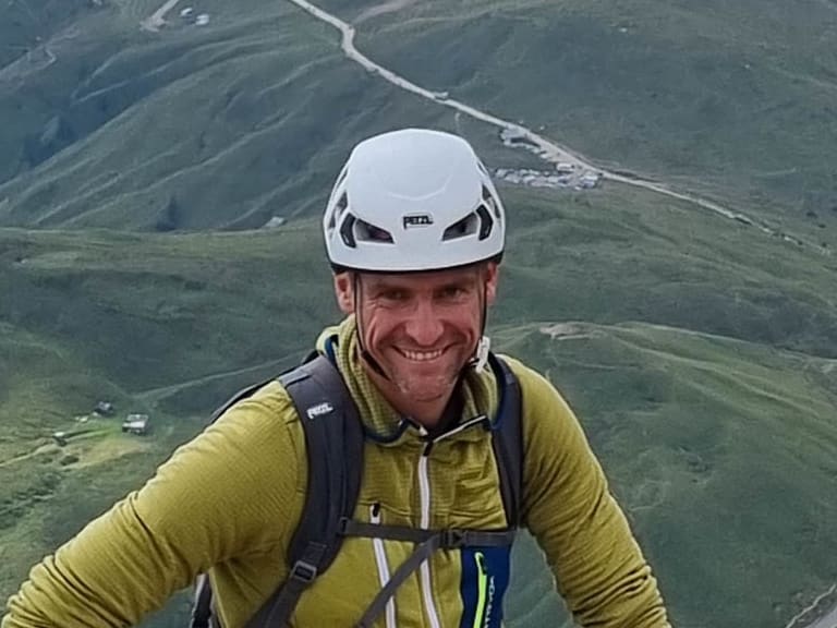 Bergführer Josef Mayr mit weißem Helm und gelbem Softshell-Oberteil lächelt am Felsgipfel mit Blick ins grüne Tal.