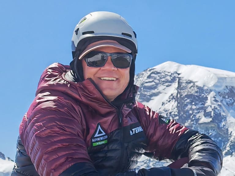 Bergführer Wolfgang Pusch sitzt in bordeauxroter Daunenjacke und weißem Helm vor einem schneebedeckten Viertausender.