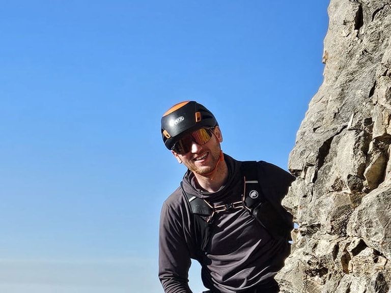 An hoher Bergwand steht Bergführer Lukas Degen mit Klettergurt und Seil vor weitem Alpenpanorama.