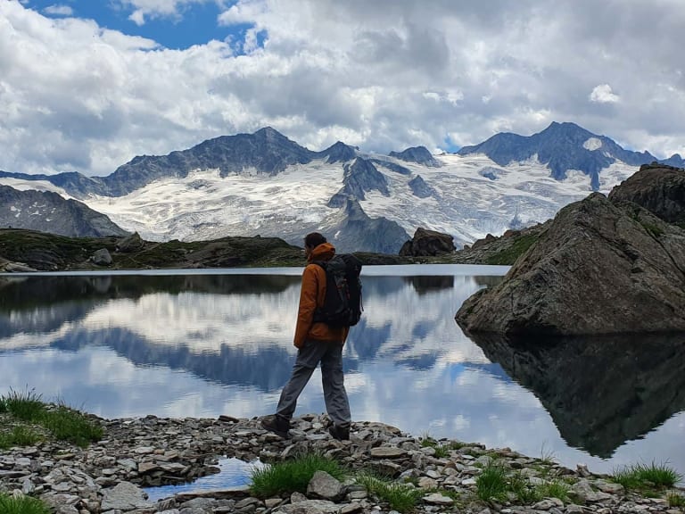 Bergwanderführer Jonas Bergmann in oranger Jacke steht am steinigen Ufer eines Bergsees vor schneebedeckter Gebirgskette.