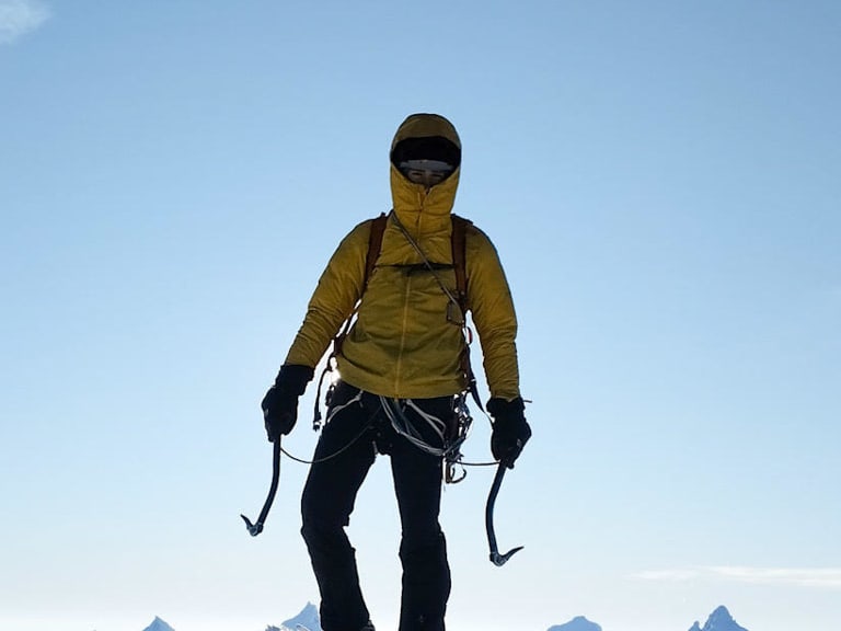 Bergführer Alexander Brückler steht auf verschneitem Berggrat mit zwei Eisgeräten in gelber Daunenjacke vor schneebedeckten Gipfeln.