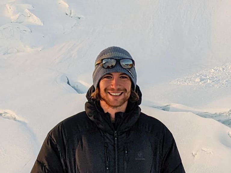 Bergführer Alexander Brückler lächelt vor verschneiter Gletscherlandschaft und trägt schwarze Daunenjacke mit grauer Mütze.