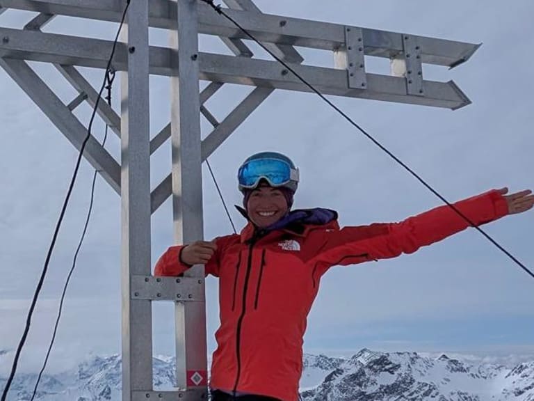 Bergführerin Monika Hümmer in roter Winterjacke mit ausgebreiteten Armen vor Holzgipfelkreuz.
