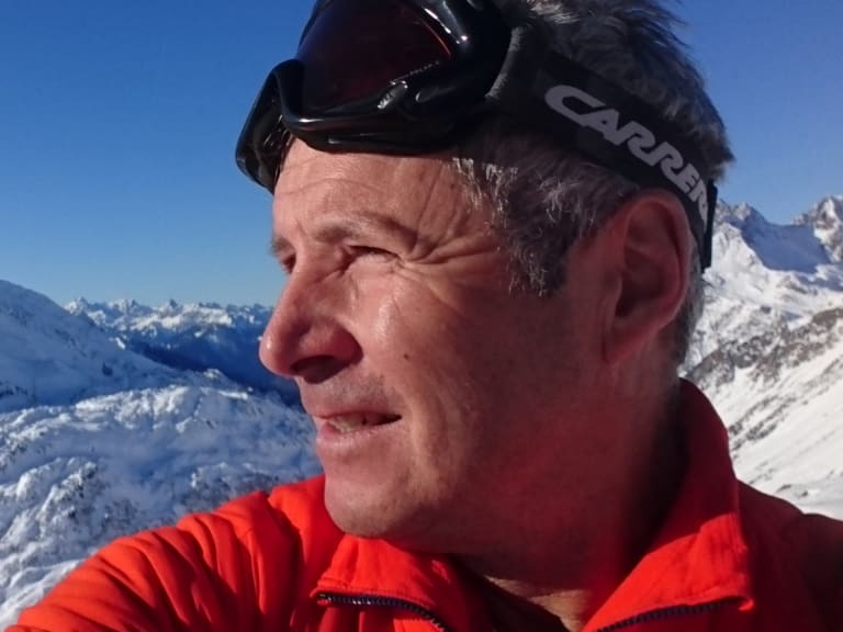 In roter Softshell-Jacke und mit Skibrille posiert Bergführer Jürgen Bäumler vor verschneiten Gipfeln.