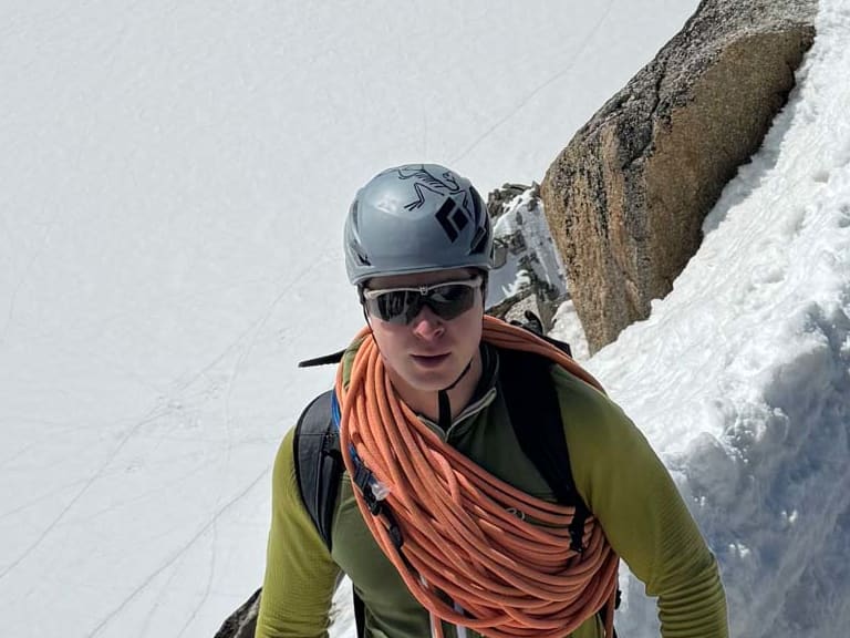 Bergführer Wolfgang Wagner steht mit orangem Kletterseil und grauem Helm auf schneebedecktem Grat in hochalpinem Gelände.