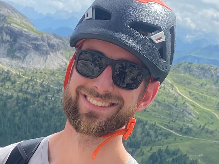 Portrait von Bergführer Joshua Wontroba mit schwarzem Kletterhelm vor weitläufiger Berglandschaft.