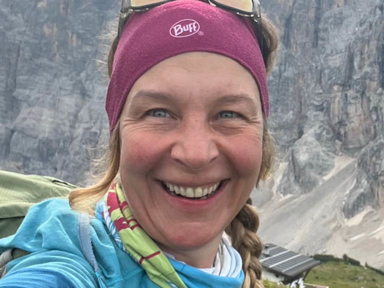 Selfie von Bergwanderführerin Bernadette Schnabl mit blonden Zöpfen, rotem Stirnband und blauer Jacke vor alpiner Felswand.