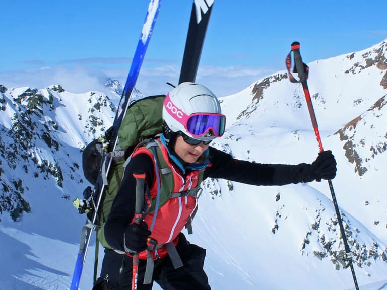 Bergwanderführerin Bernadette Schnabl steigt auf Skiern durch Pulverschnee auf, trägt weißen Helm und rot-schwarze Jacke vor Schneegipfeln.