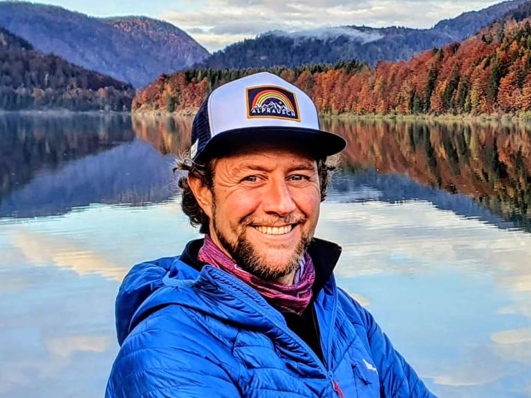 Am ruhigen Bergsee mit herbstlicher Bergkulisse steht Bergwanderführer Stephan Wild in blauer Daunenjacke und Baseballkappe.
