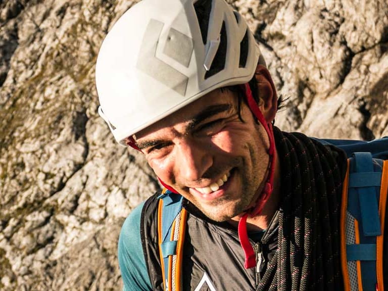 Bergführer Josef Neumeier mit weißem Helm und blauer Kleidung lächelt vor einer steilen Kalksteinfelswand.