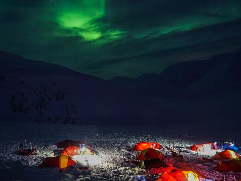 Bergwanderführer Jonas Haass steht bei beleuchteten Expeditionszelten auf schneebedecktem Terrain unter grünen Nordlicht-Schleiern am Nachthimmel.