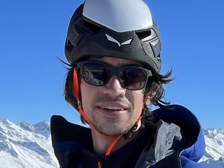 Bergführer Michael Gruber trägt blaue Skijacke und weißen Helm, neben ihm ist die Spitze eines orange-schwarzen Tourenskis sichtbar.