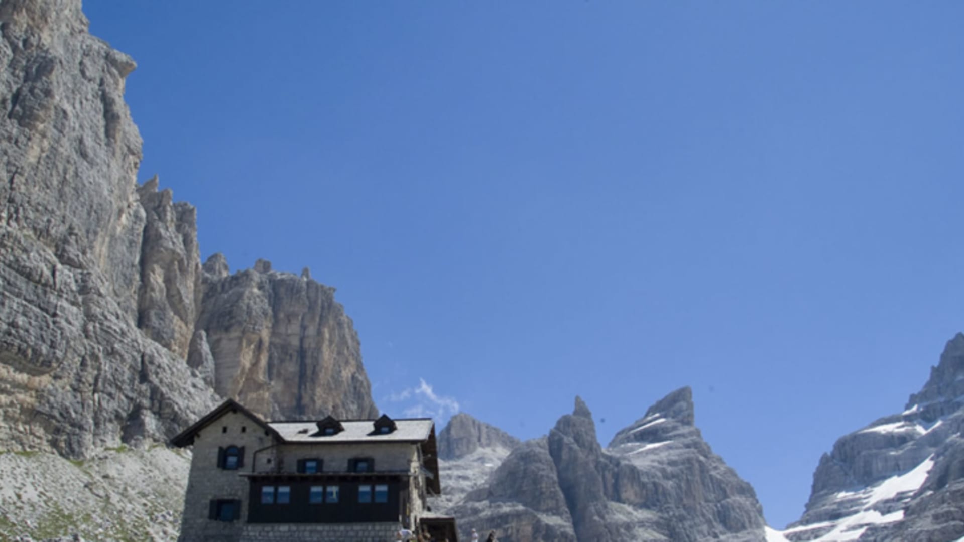 Das Rifugio Tuckett steht auf einem Felsplateau vor steilen Felswänden mit zwei Bergwanderern davor.