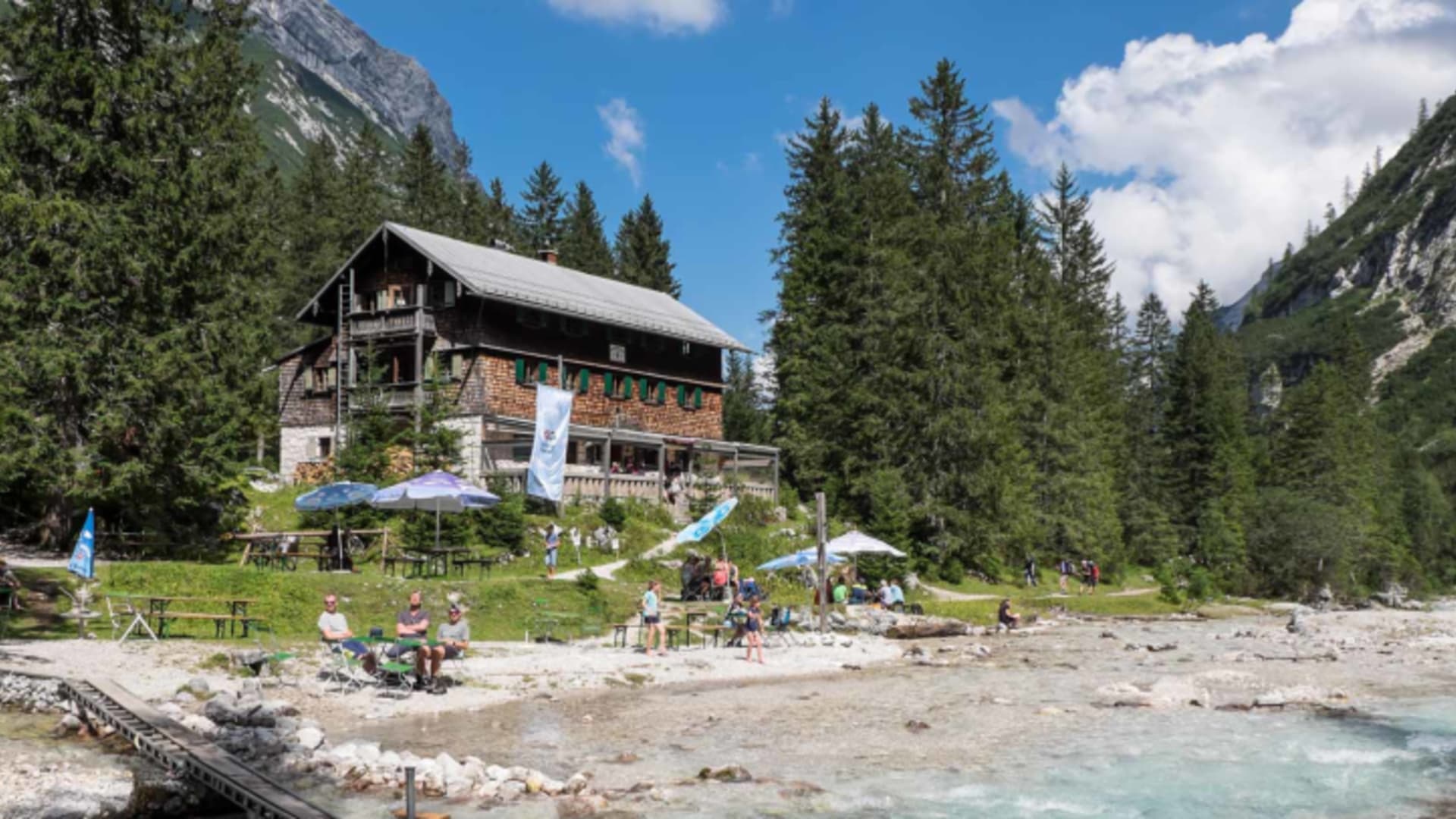 Panoramaaufnahme der Reintalangerhütte mit Holzsteg am türkisblauen Bergsee und Gästen unter Sonnenschirmen.