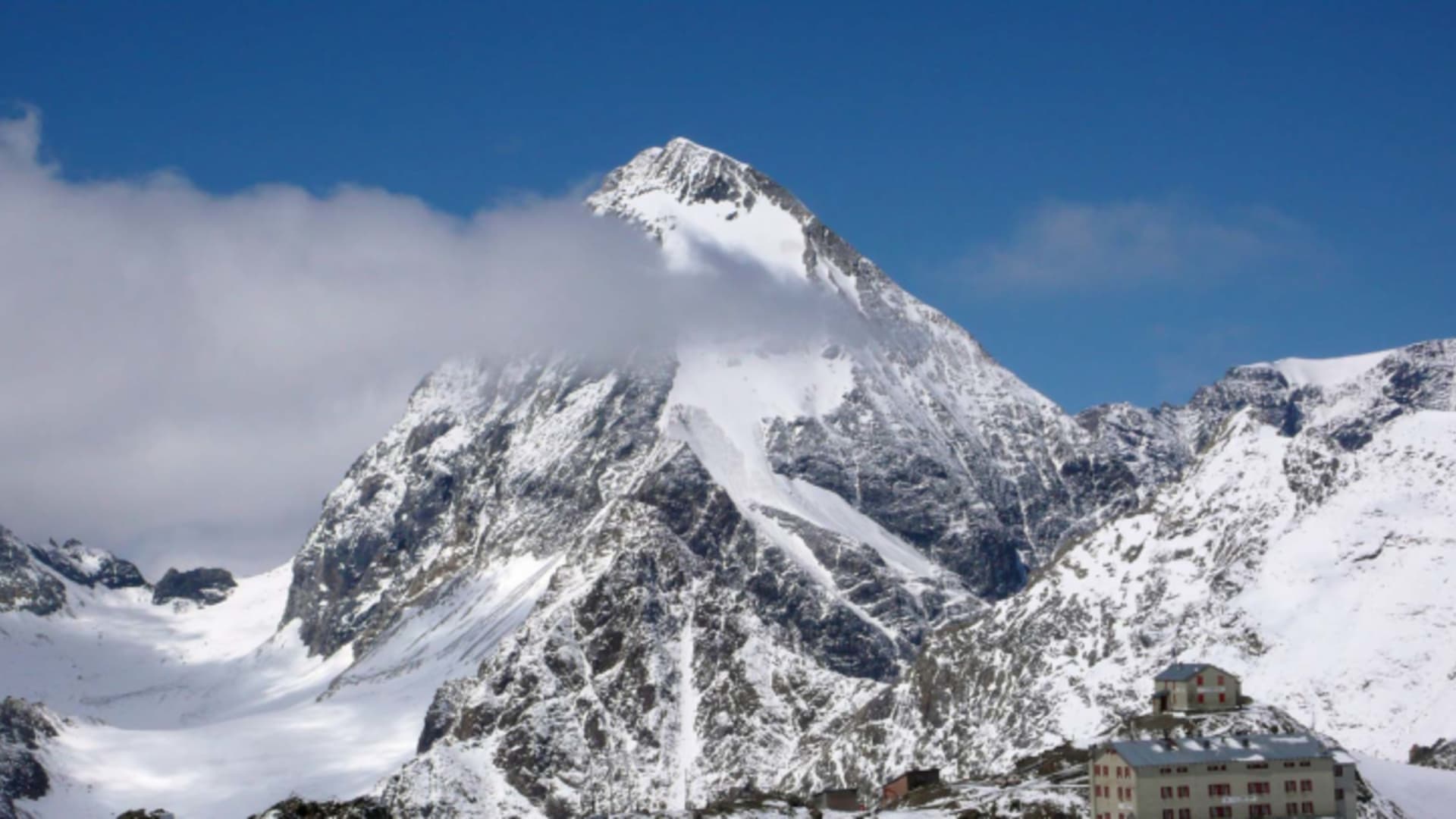Panorama des Rifugio Casati mit rosa-rötlicher Fassade auf Felssockel umgeben von Gletschern und verschneiten Gipfeln.