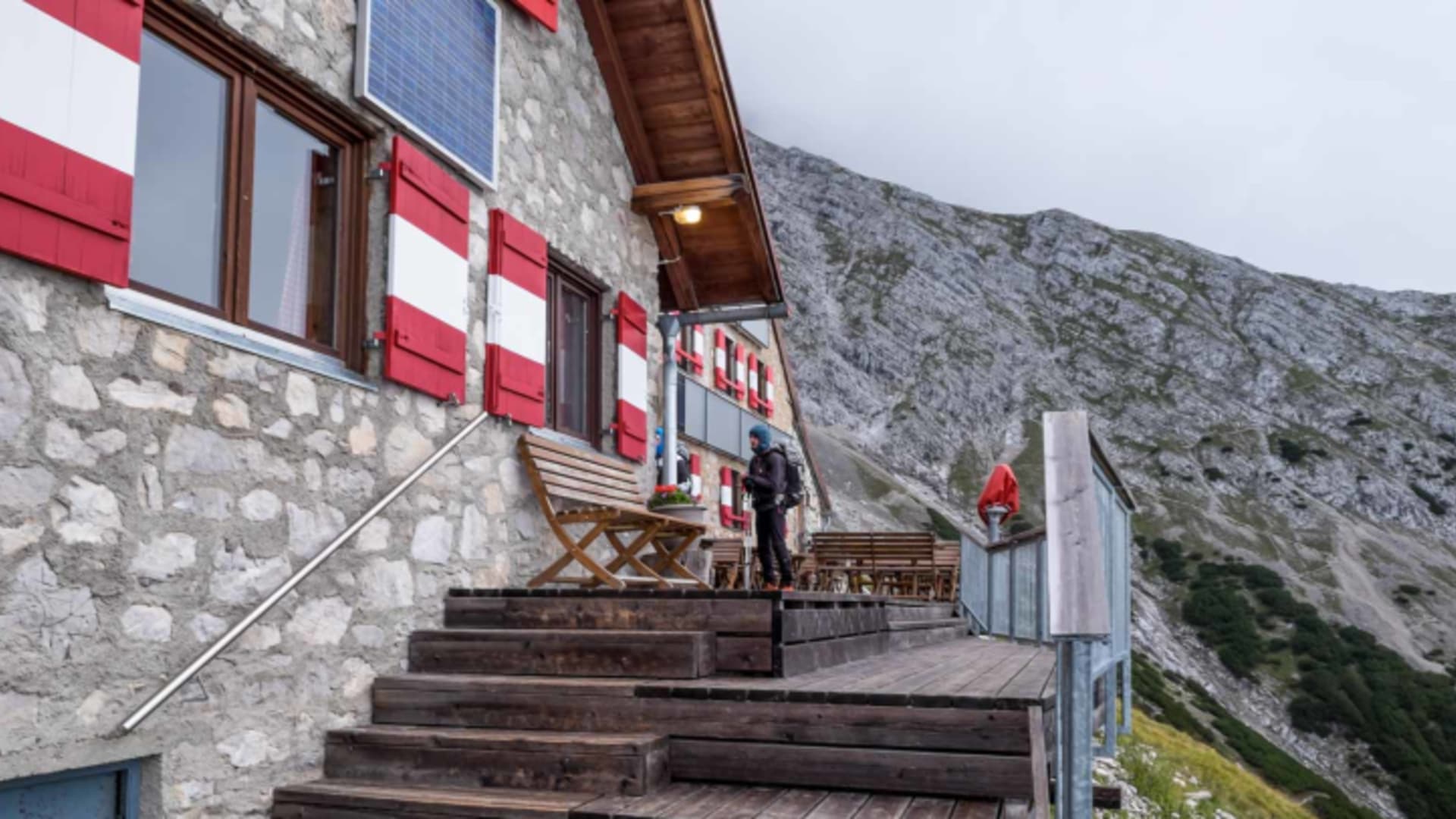 Außenansicht der Bettelwurfhütte mit Natursteinmauern, Holzterrasse und Wanderern vor dem Eingang.