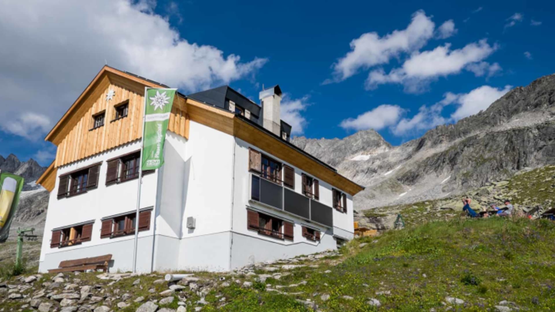 Wanderer bei der Plauener Hütte mit grüner Edelweiss-Fahne vor Bergkulisse.