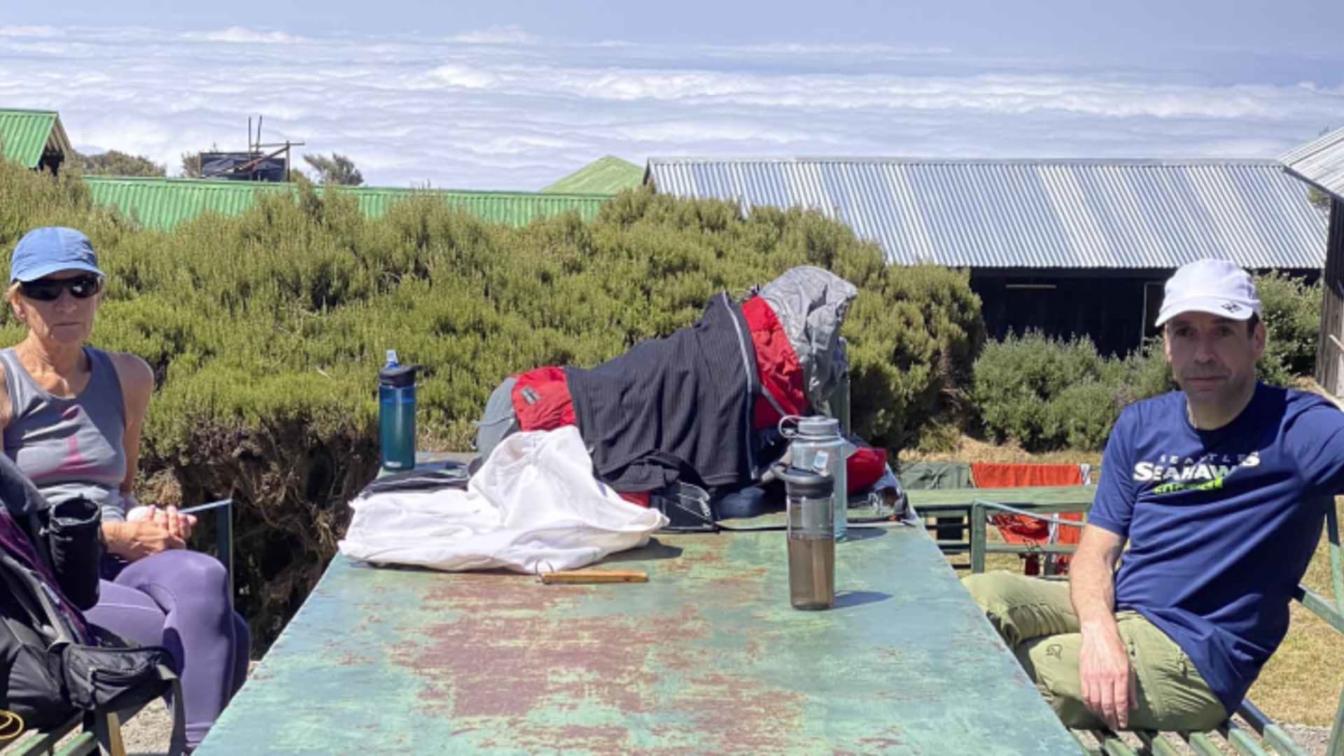 Zwei Bergsteiger pausieren an der Sattelhütte (Mt. Meru) vor Wellblechhütten mit Bergpanorama.