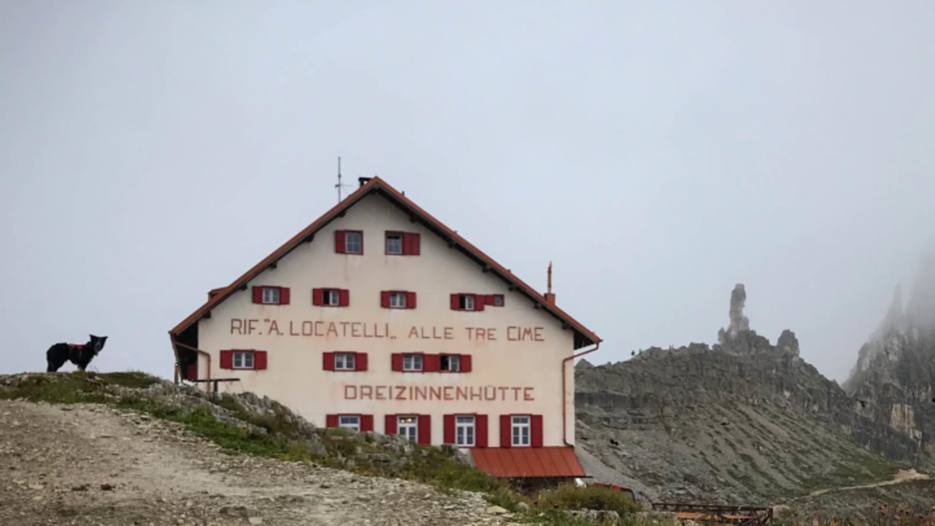 Die Drei Zinnen Hütte mit weißer Fassade und rotem Dach steht auf steinigem Plateau unter Dolomitengipfeln.