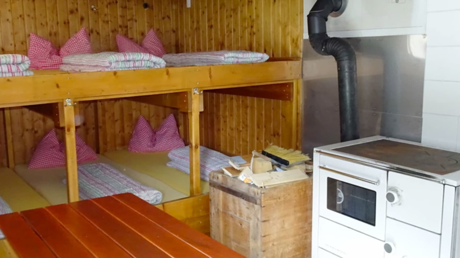 Im Westfalenhaus zeigt der Winterraum vier Stockbetten aus Holz mit rot-weißen Kissen und einen traditionellen Küchenofen.