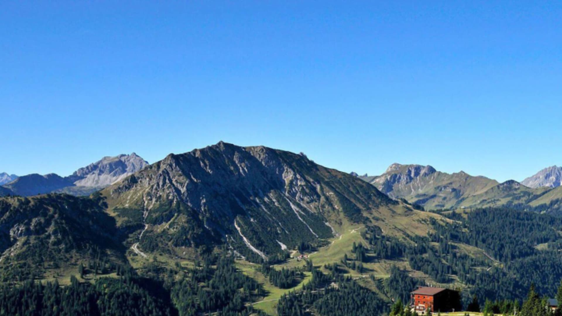 Das Gimpelhaus mit roter Holzfassade steht auf einem bewaldeten Hang mit weitem Panoramablick über die alpine Berglandschaft.