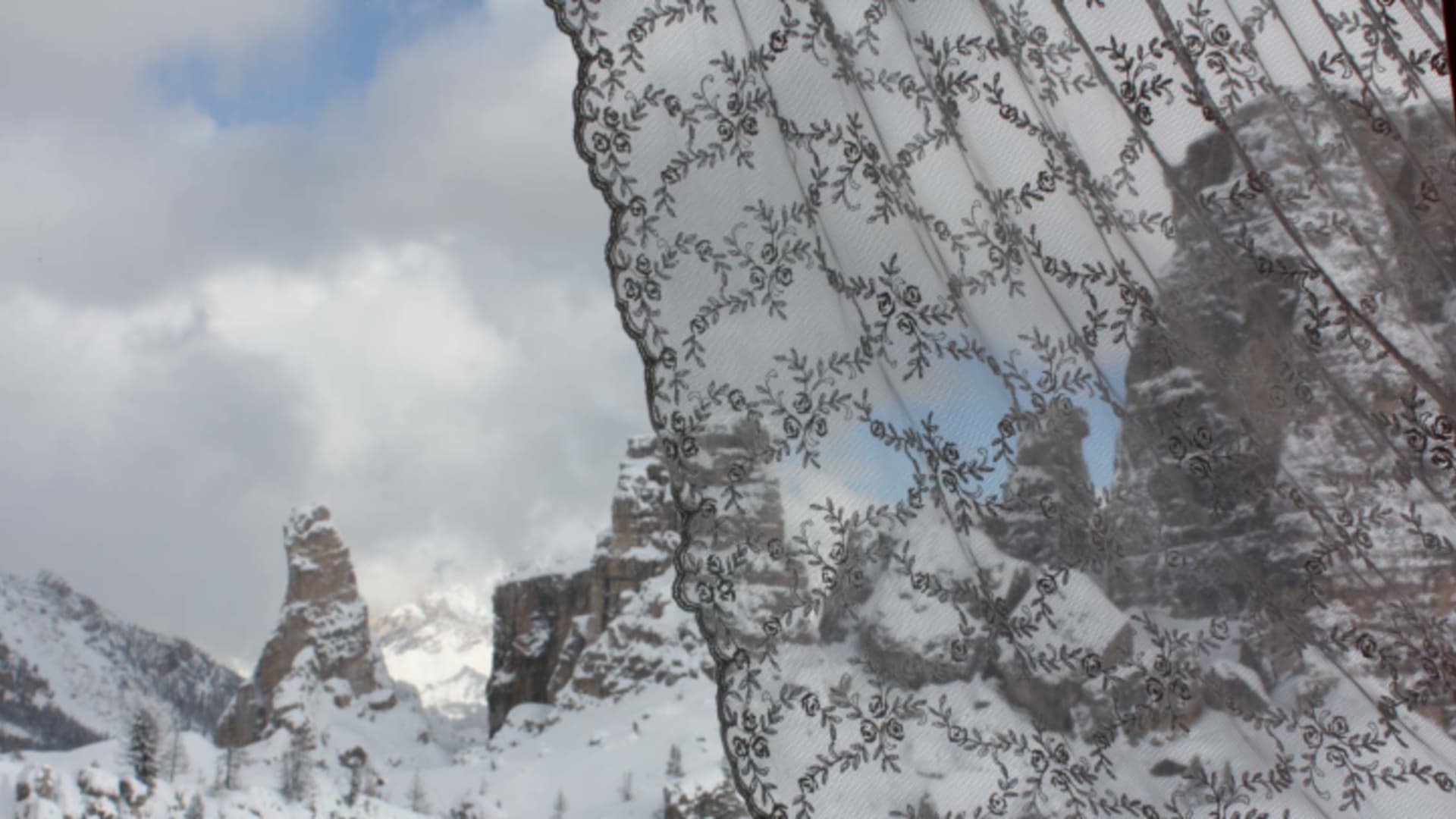 Blick durch das Fenster des Rifugio Scoiattoli auf verschneite Dolomiten-Felswände hinter Spitzenvorhang.