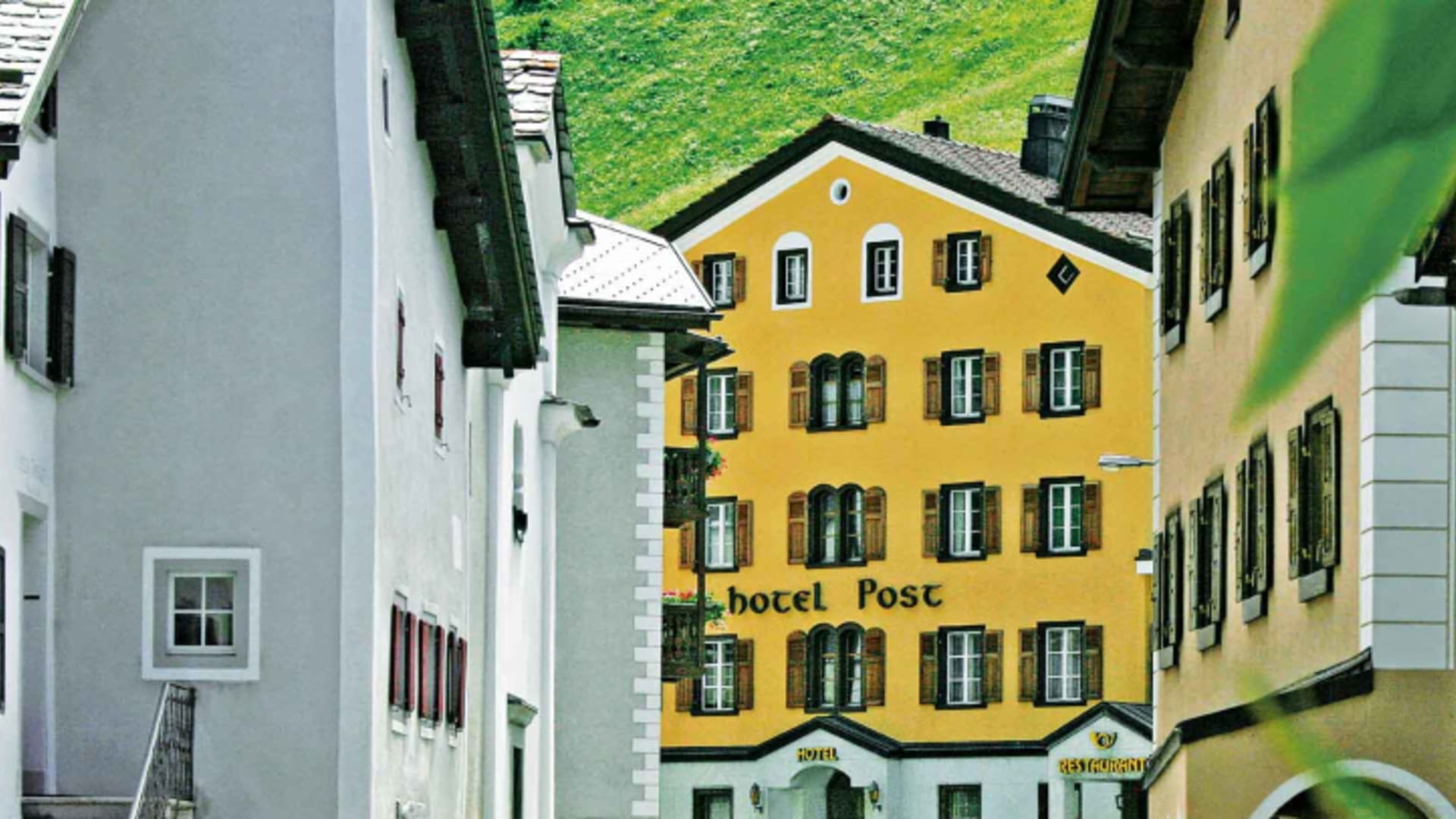 Sommerliche Außenansicht des Hotel Post Bivio mit gelber Fassade inmitten des Bergdorfs in grüner Alpenlandschaft.