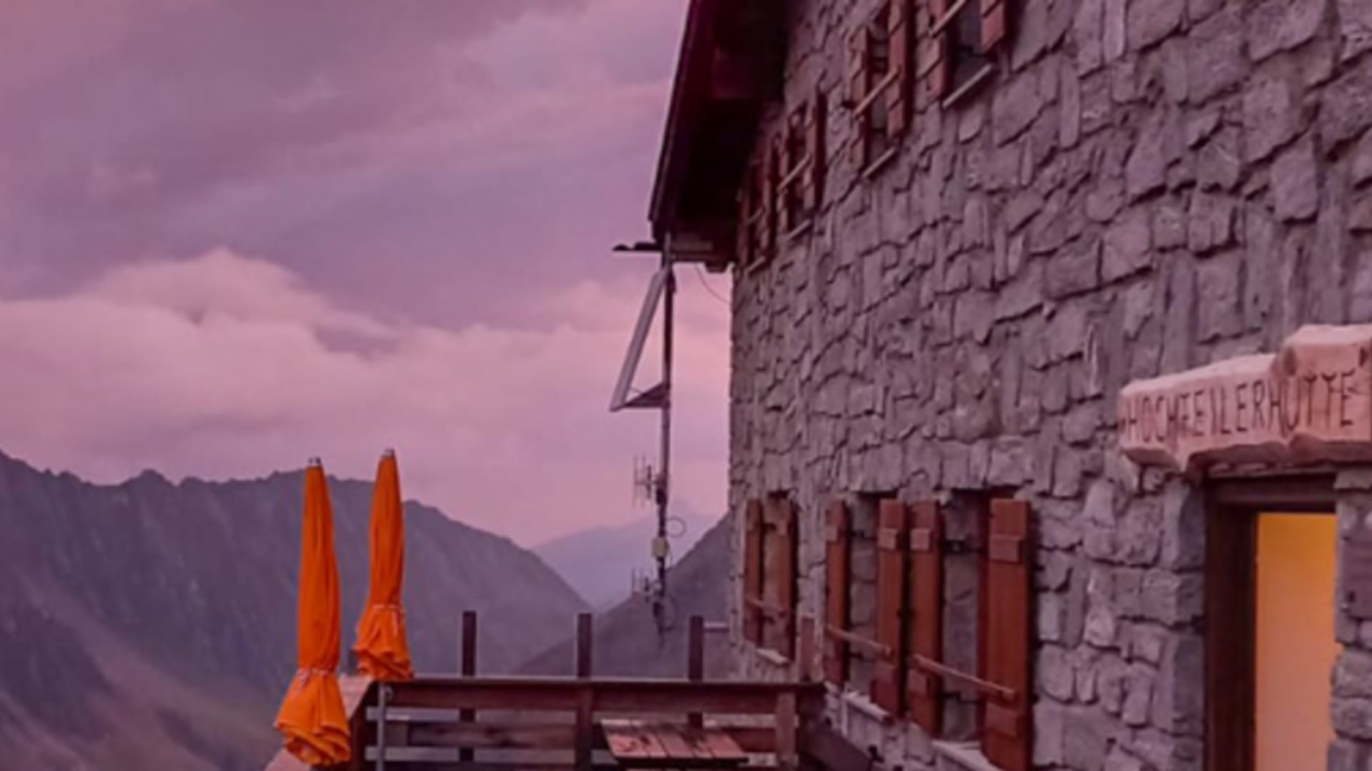 Terrasse der Hochfeilerhütte mit Holztischen und zusammengeklappten Sonnenschirmen im violetten Abendlicht.