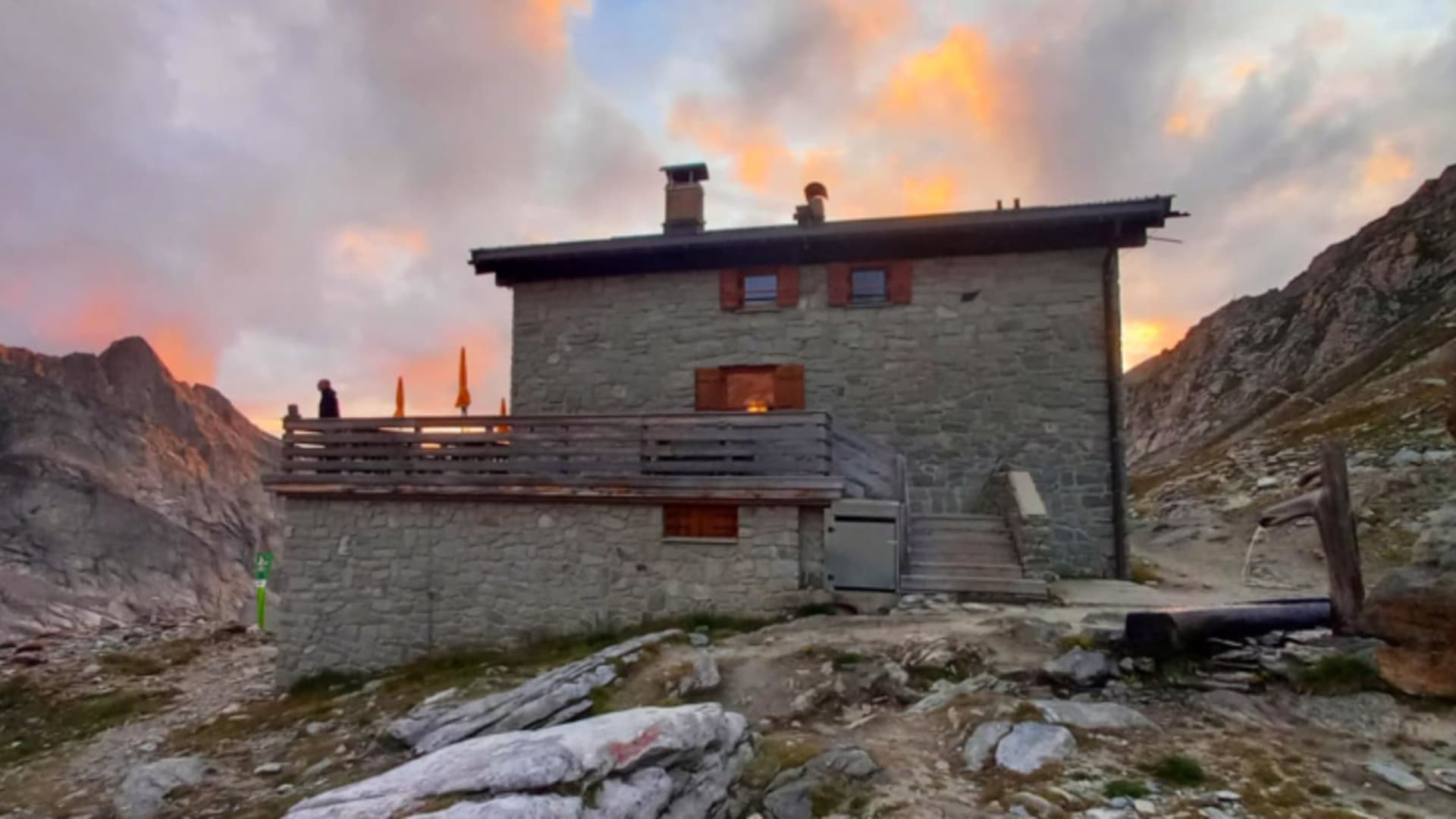 Die Hochfeilerhütte aus Naturstein mit Holzbalkon, auf dem eine Person sitzt, umgeben von steilen Felswänden.