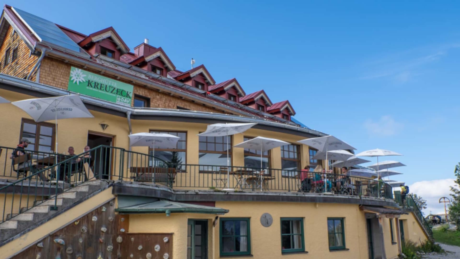 Außenansicht des Kreuzeckhauses mit gelber Fassade, großer Terrasse mit Sonnenschirmen und einer Kletterwand.