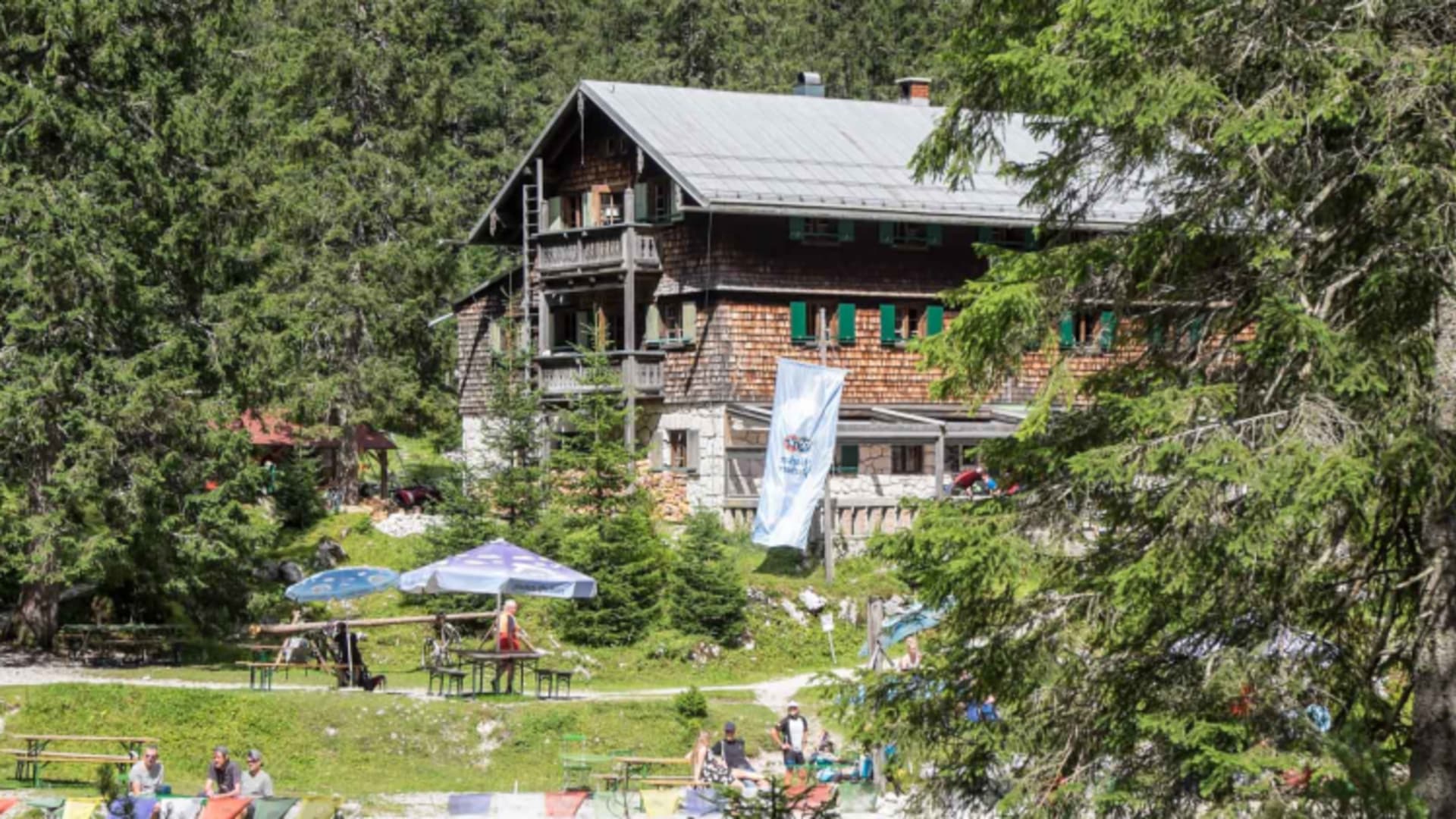 Bergsteiger entspannen auf der Terrasse der Reintalangerhütte mit orangefarbenen Liegestühlen und blauem Sonnenschirm.